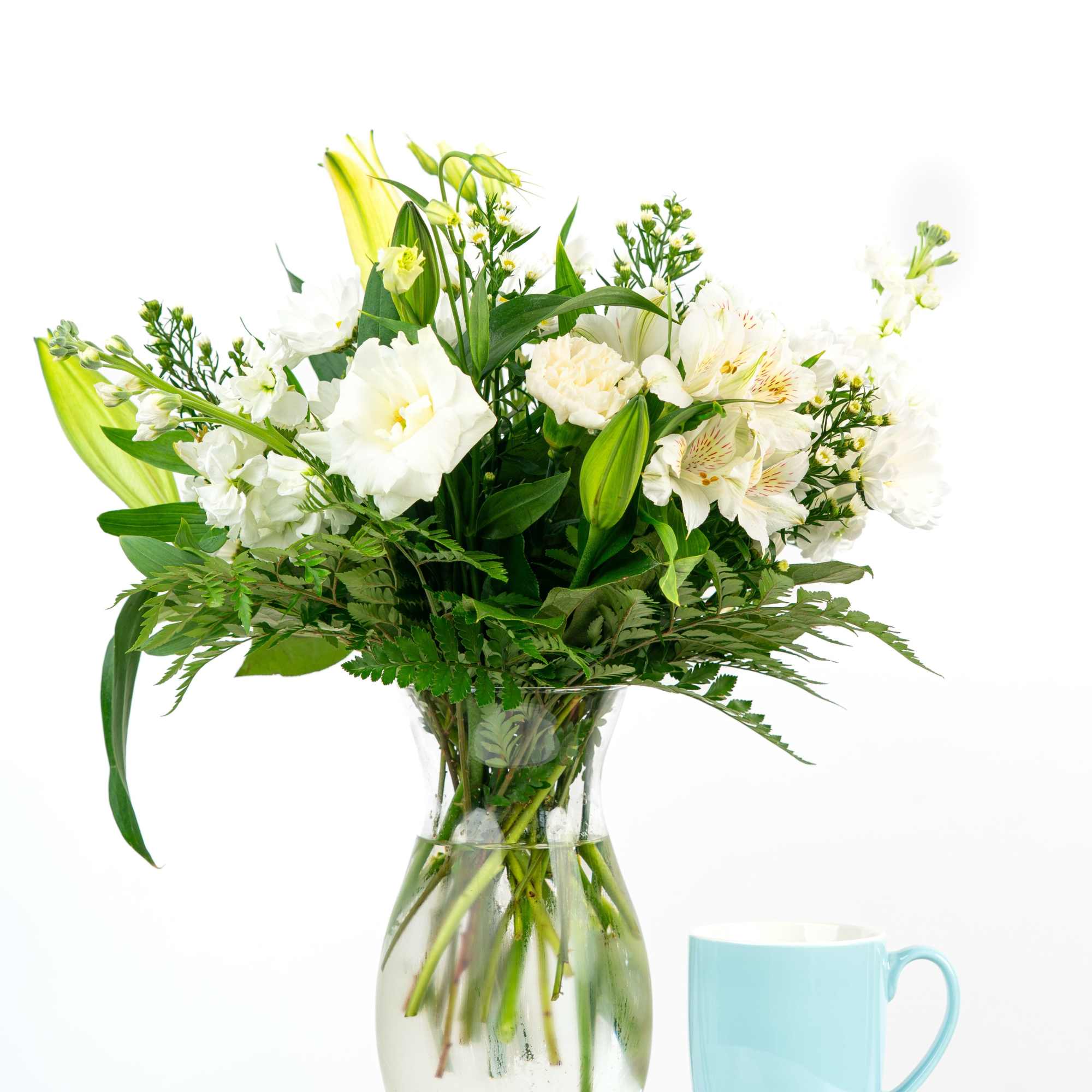 White lilies and mixed white blooms arranged in a clear glass vase beside a light blue mug.