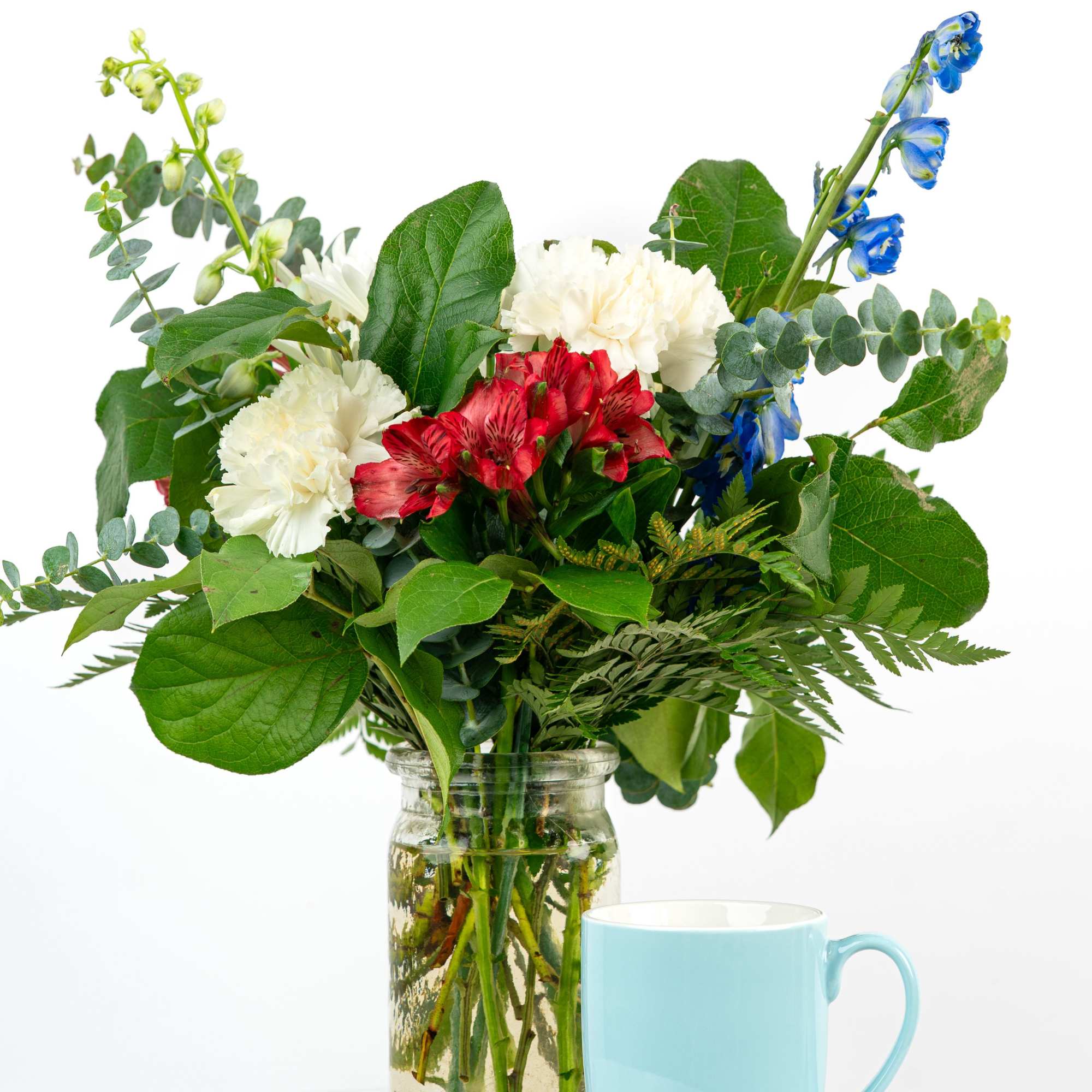 Mixed bouquet of white carnations, red alstroemeria, and blue blooms in a glass jar beside a blue mug