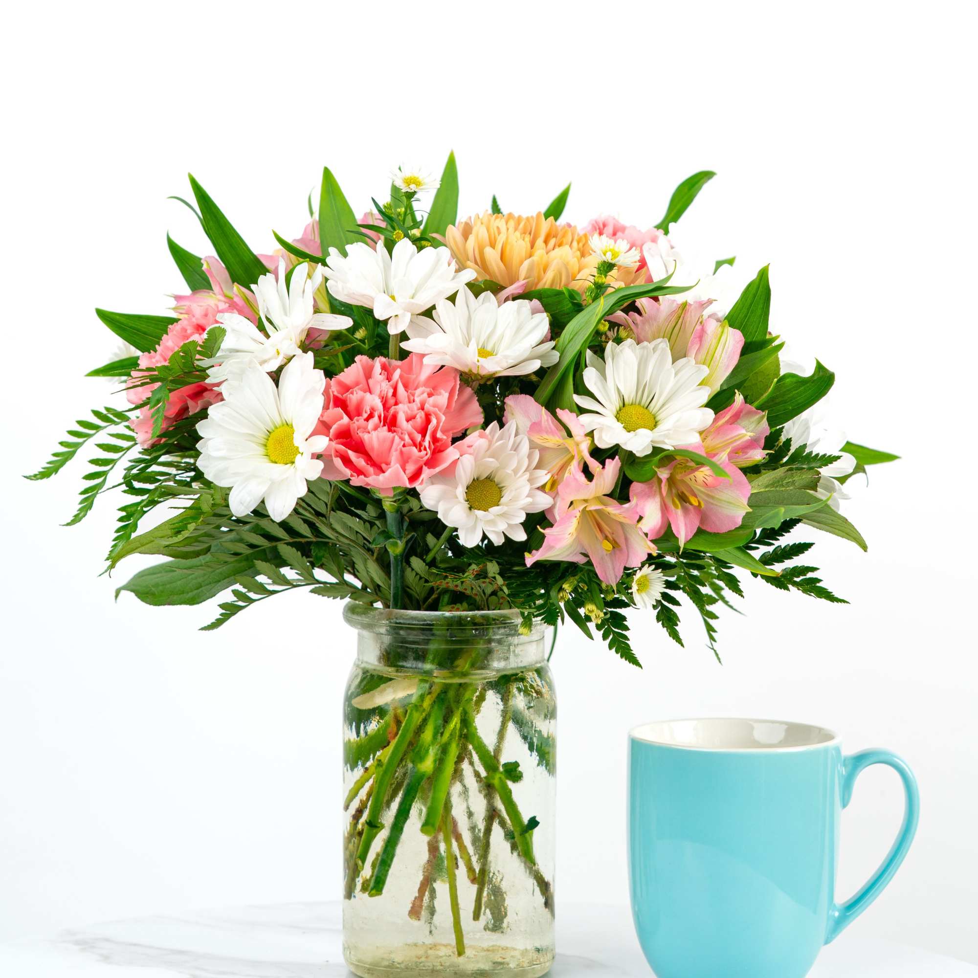 Mixed bouquet of white daisies and pink flowers in a glass jar beside a light blue mug