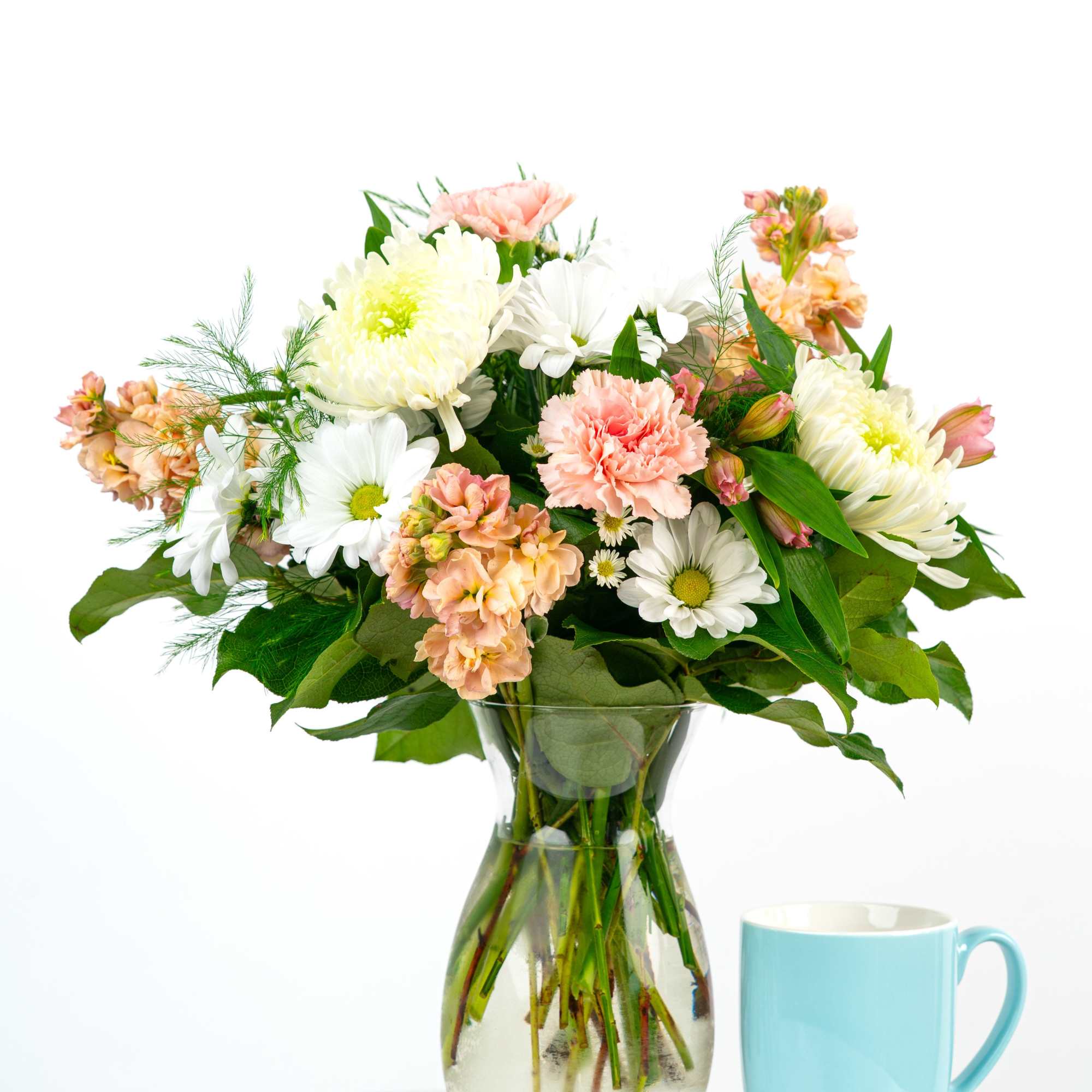 Mixed bouquet of white daisies, mums, and peach carnations in a clear vase beside a blue mug