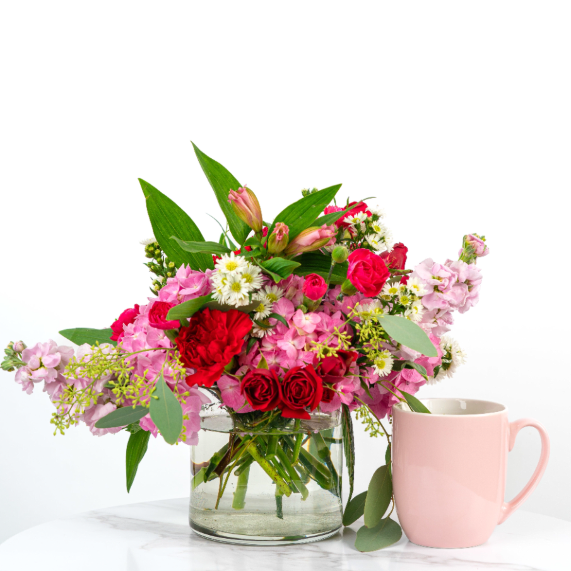 Pink and red mixed flower arrangement in a clear glass vase beside a pink mug