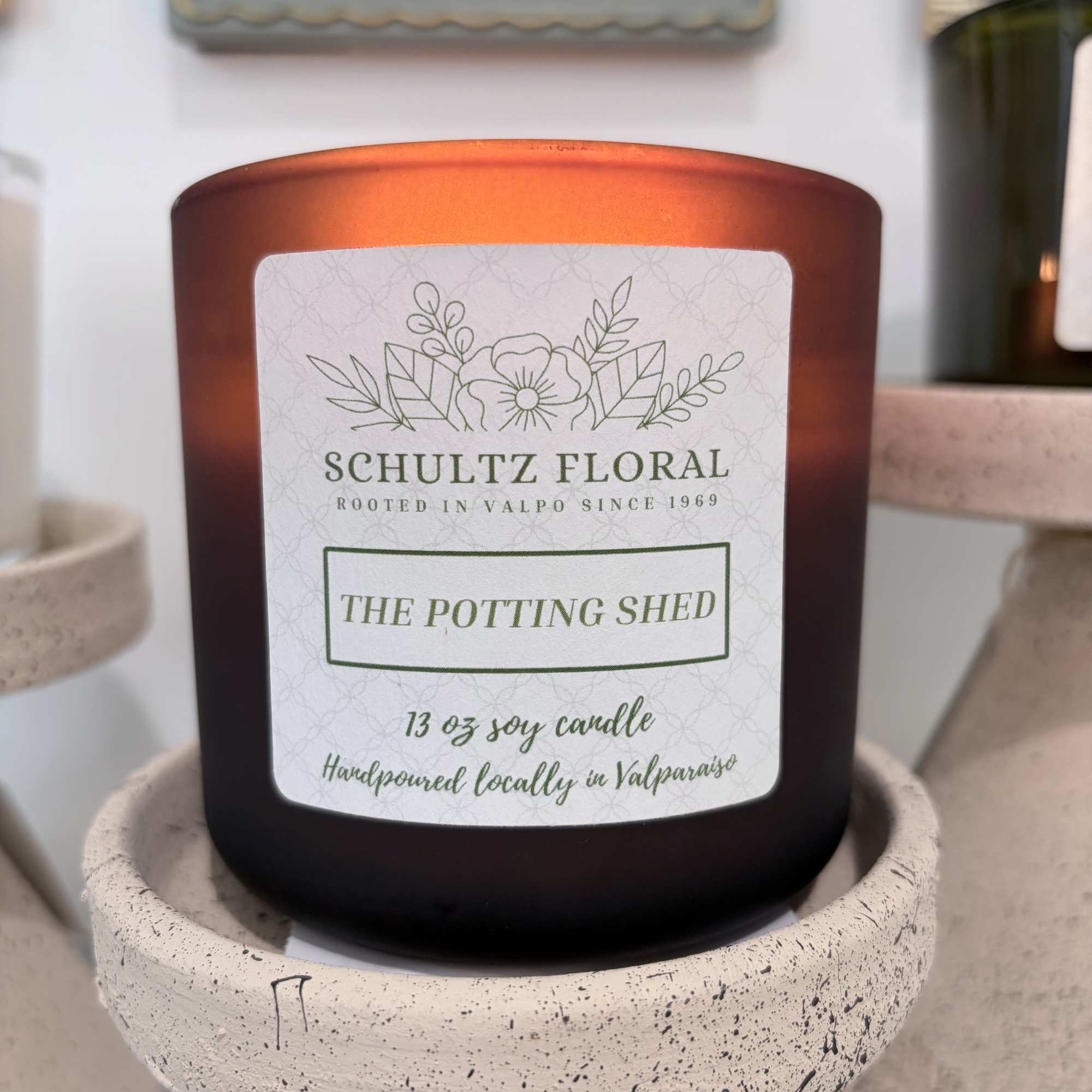 Amber glass candle labeled The Potting Shed on a speckled pedestal