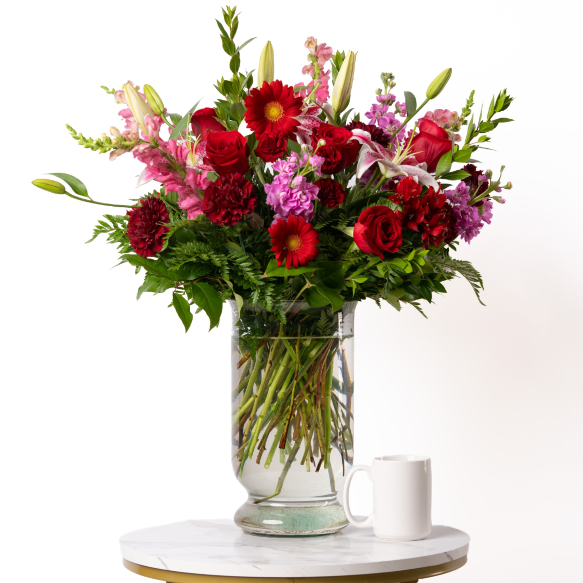 Mixed bouquet of red and pink flowers in a glass vase beside a white mug