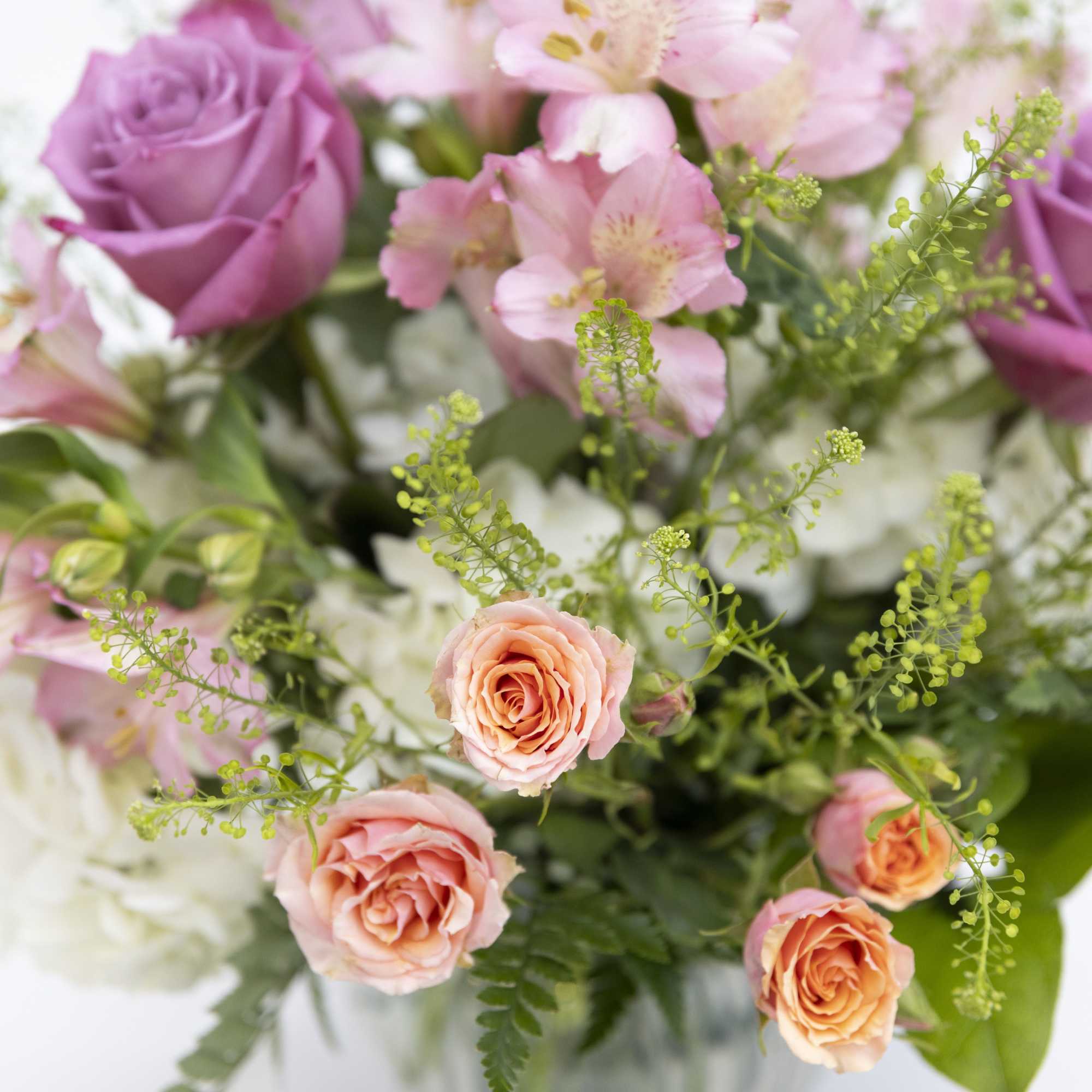 Mixed bouquet of peach spray roses, lavender roses, pink blooms, and white flowers in a clear vase