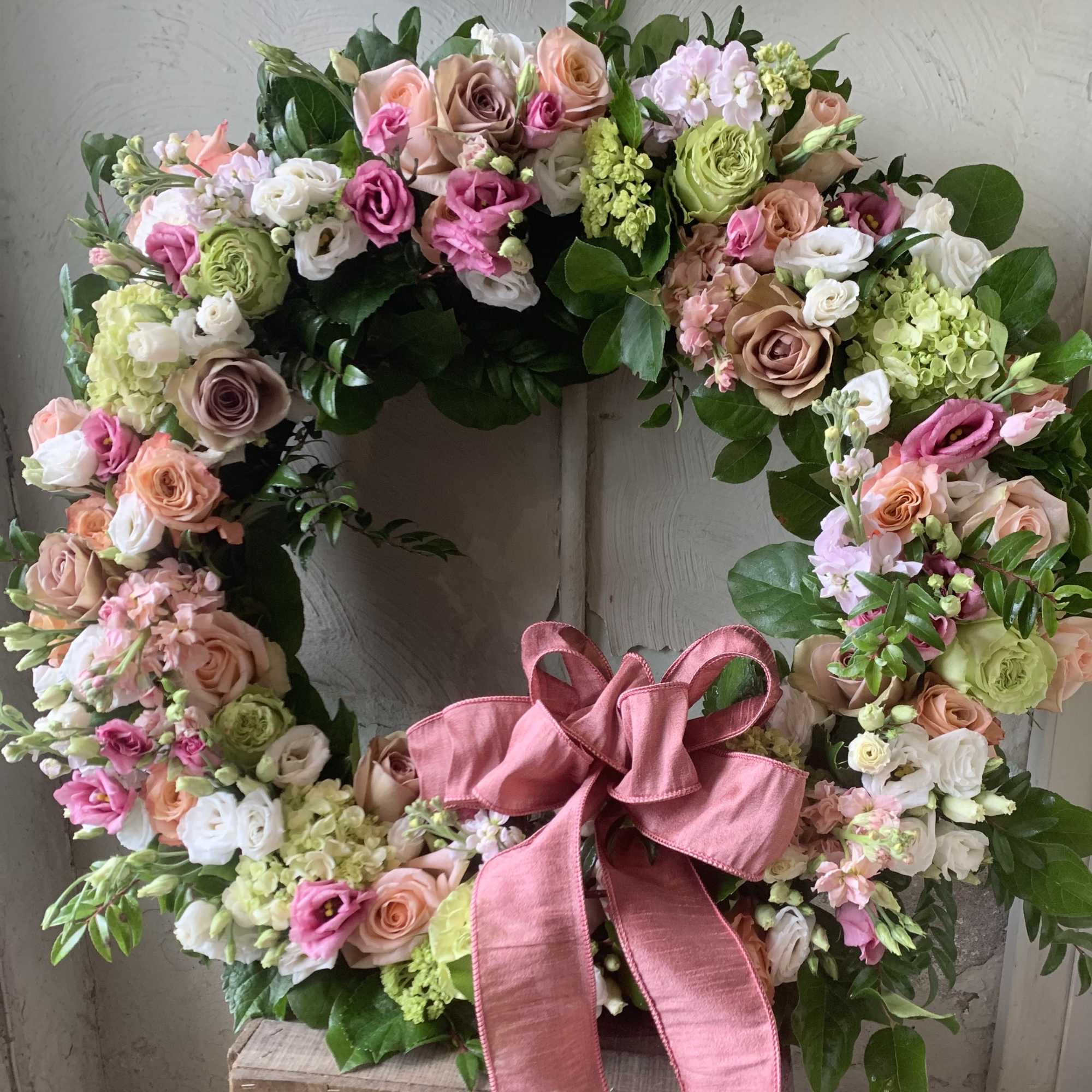 Heart-shaped floral wreath with pink, peach, white, and green blooms and a pink ribbon