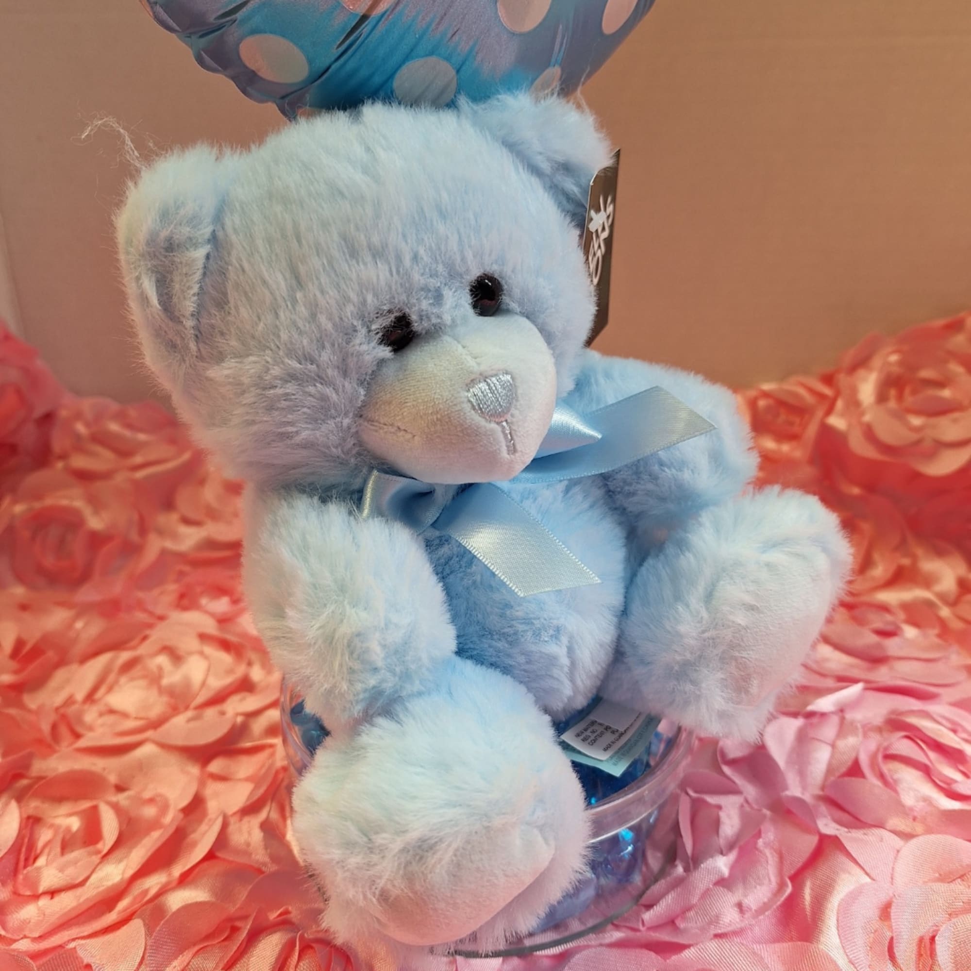 Blue teddy bear with a ribbon sitting on a clear container against pink fabric