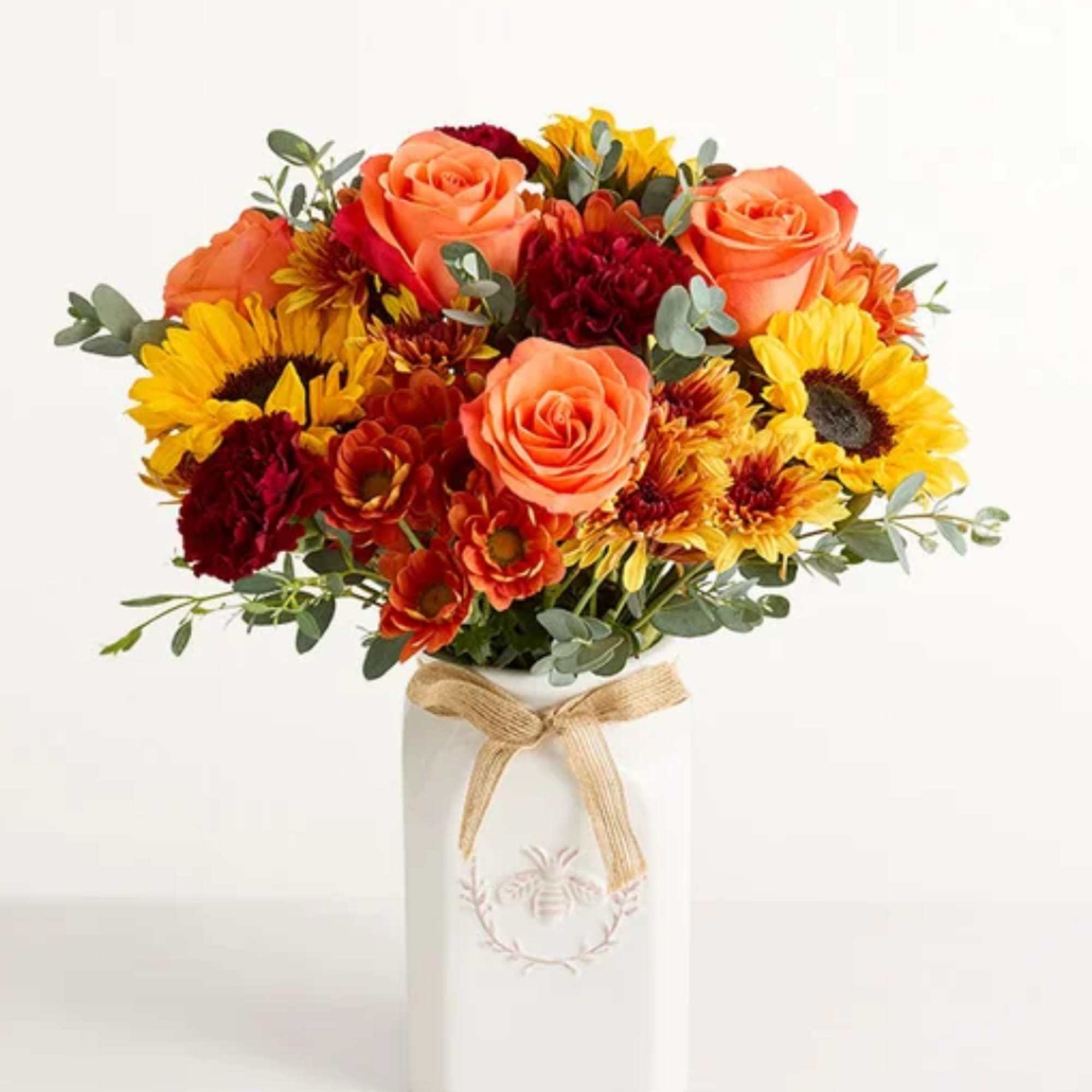 Bouquet of orange roses, sunflowers, and red blooms in a white vase with a bow