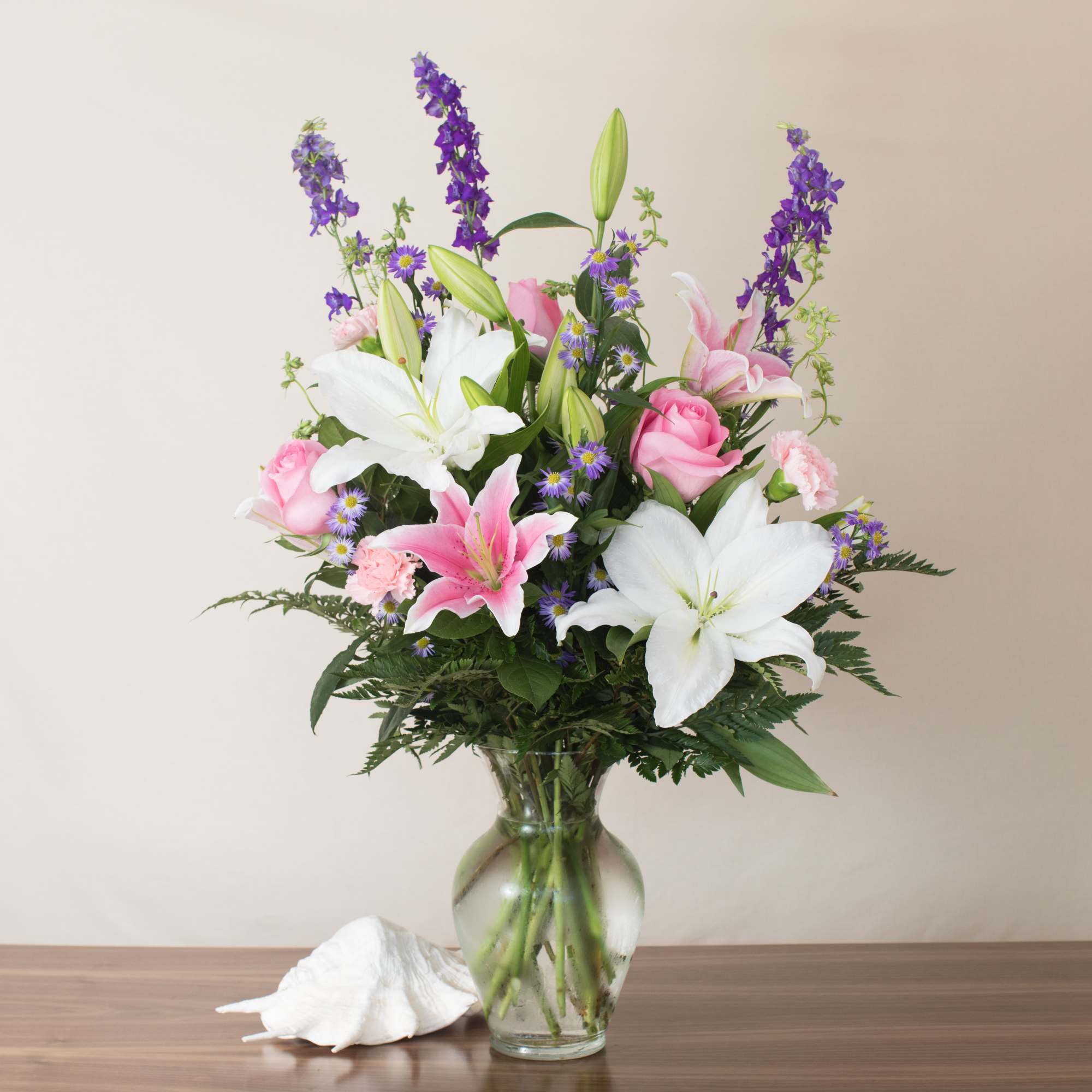 Tall arrangement of white lilies, pink roses, and purple flowers in a clear glass vase.