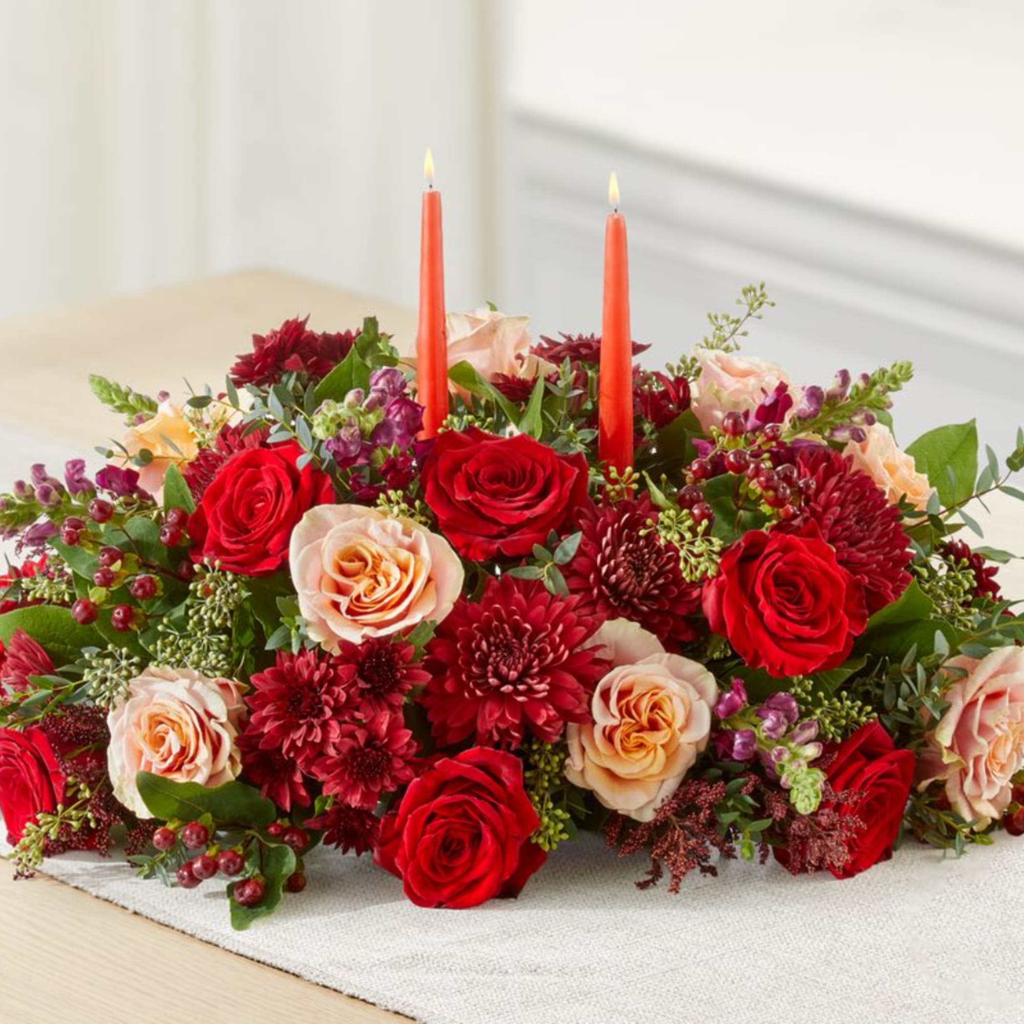 Red and peach roses with burgundy blooms and two orange candles in a low centerpiece