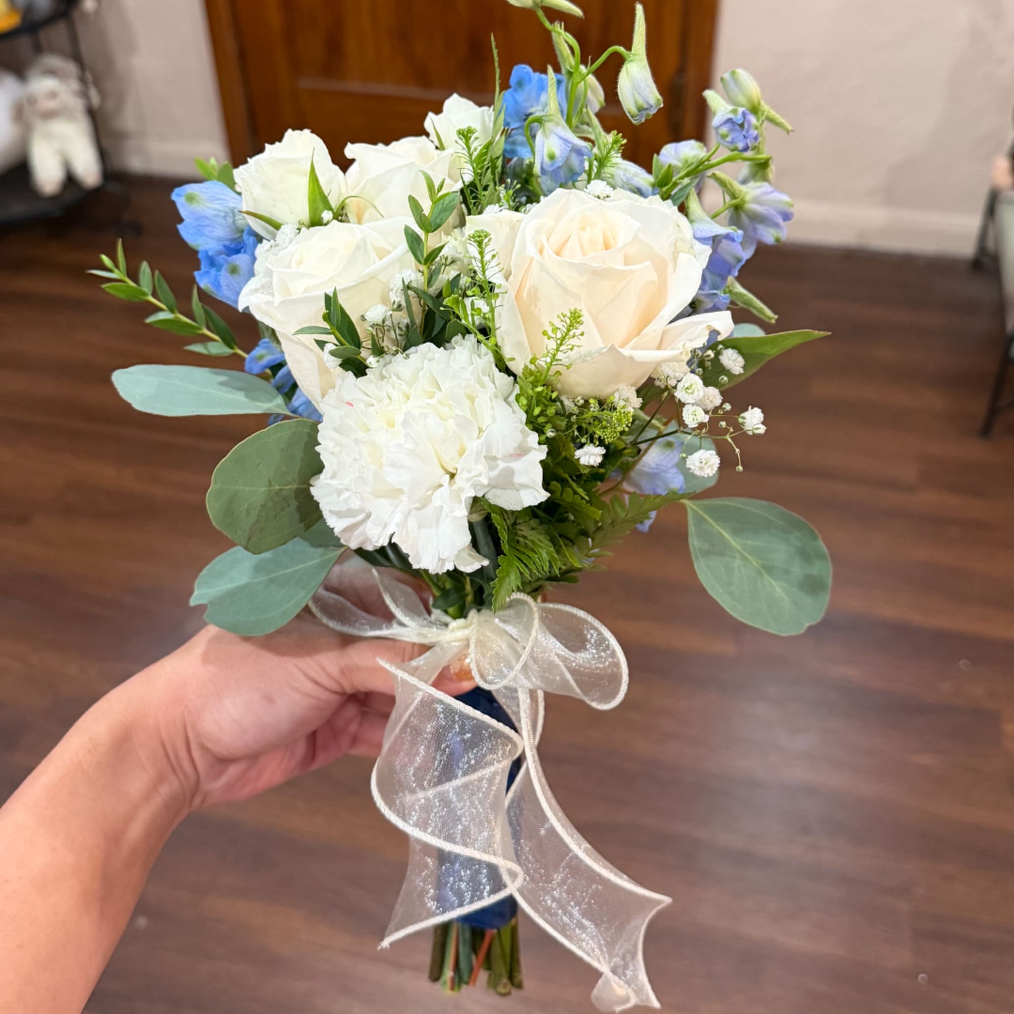 Handheld bouquet of white roses, white carnations, and blue flowers tied with a sheer ribbon