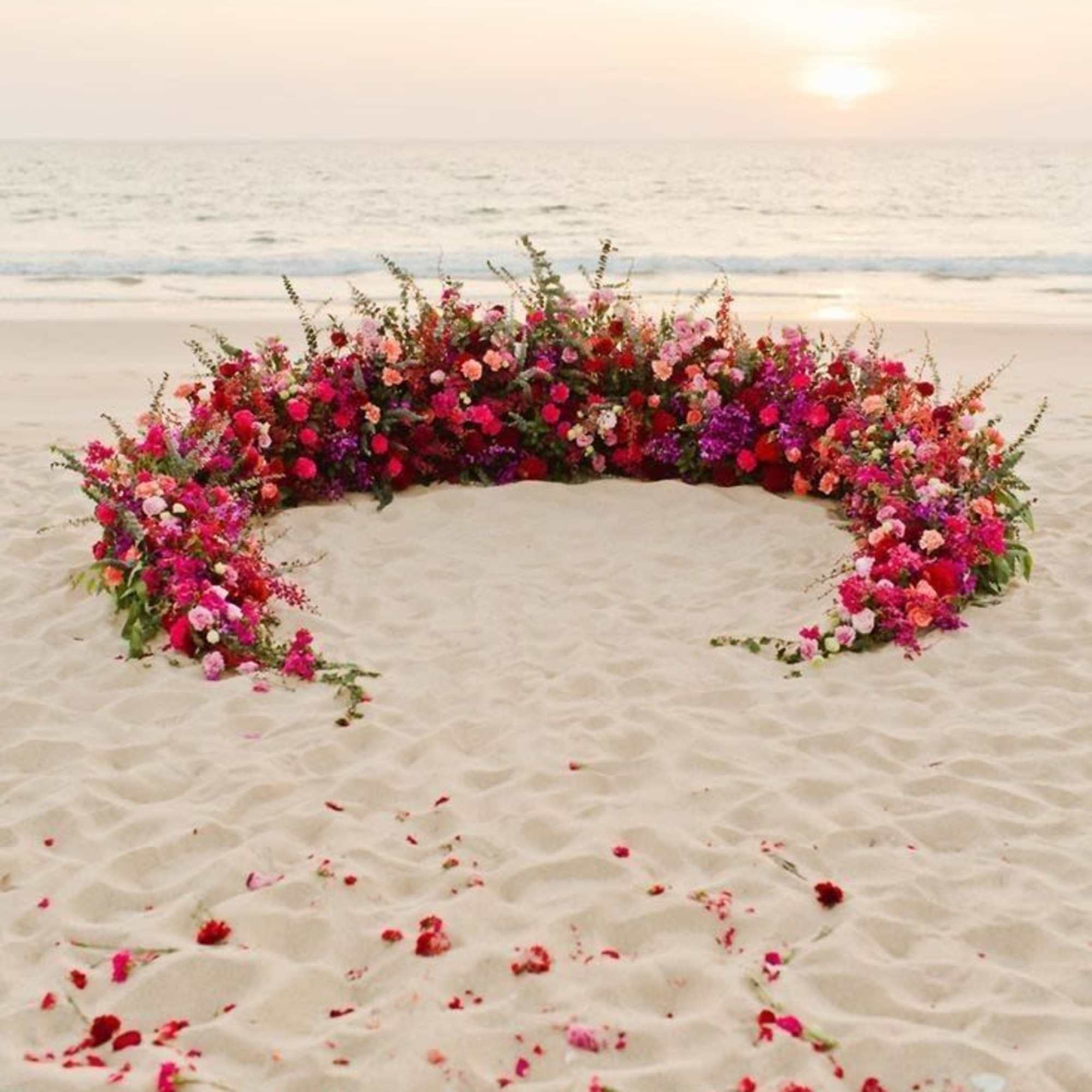 Large crescent-shaped floral arrangement on a beach at sunset