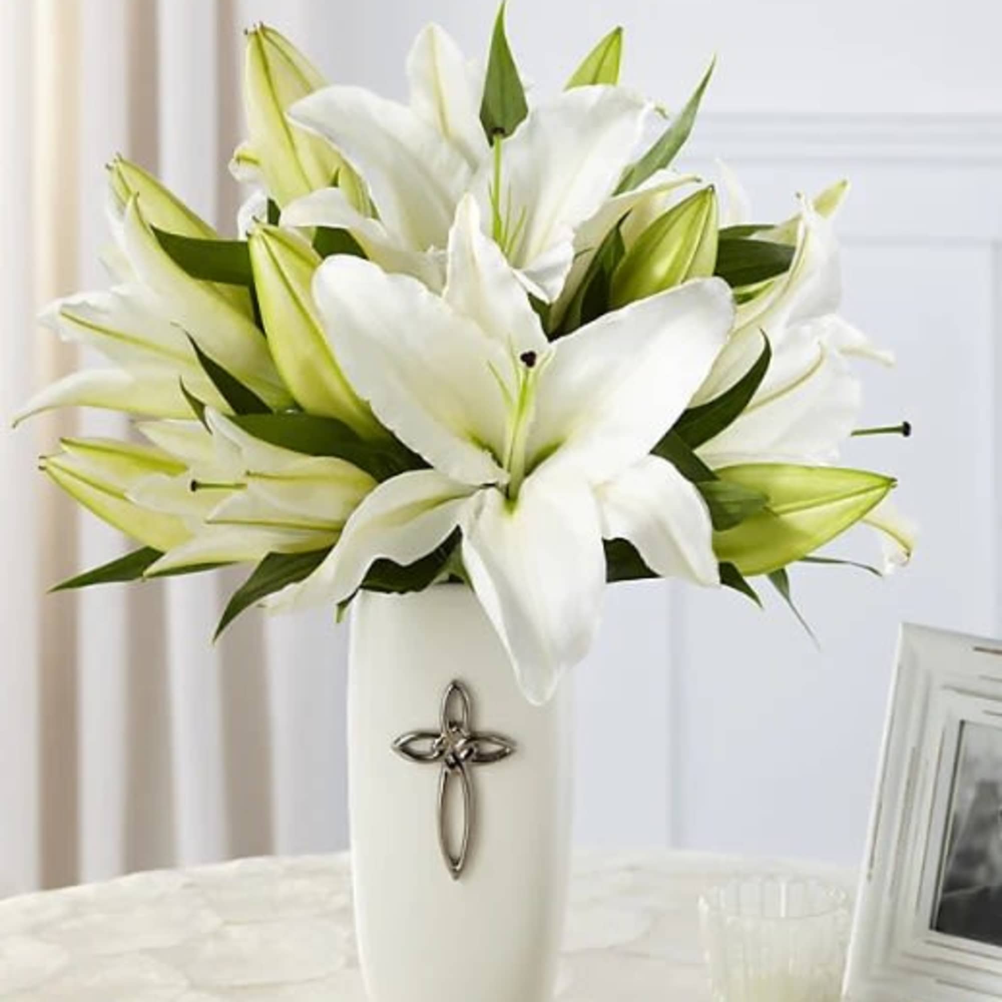 White lilies arranged in a tall white vase with a cross emblem