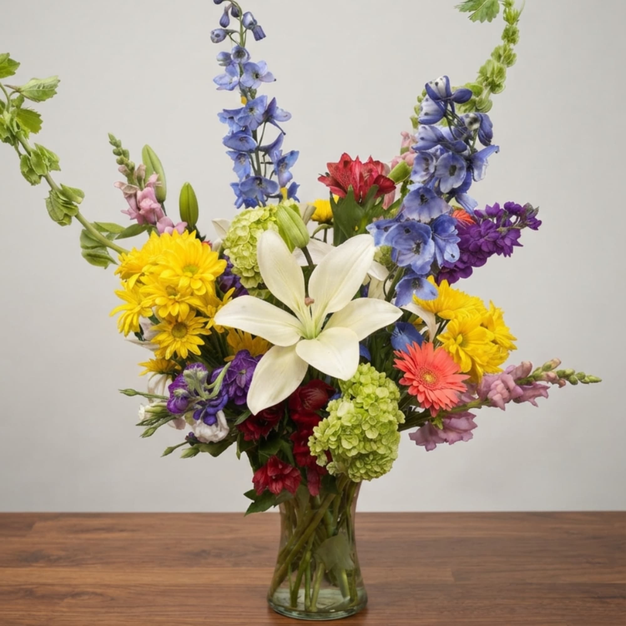Colorful mixed bouquet with lilies, daisies, and tall blue flowers in a glass vase