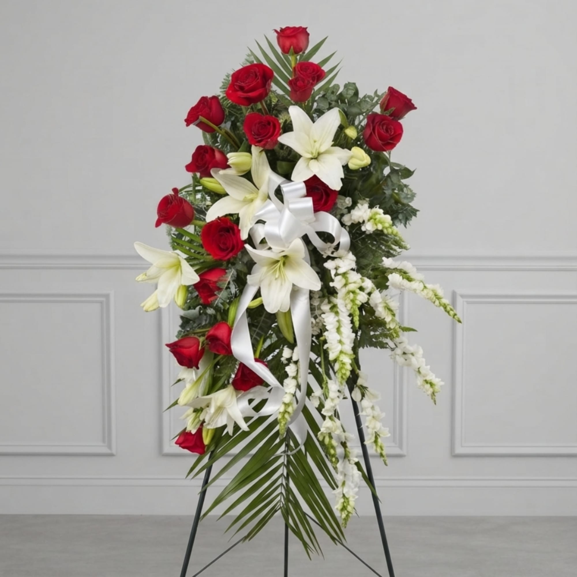 Standing floral spray with red roses, white lilies, and a white ribbon on a metal easel.