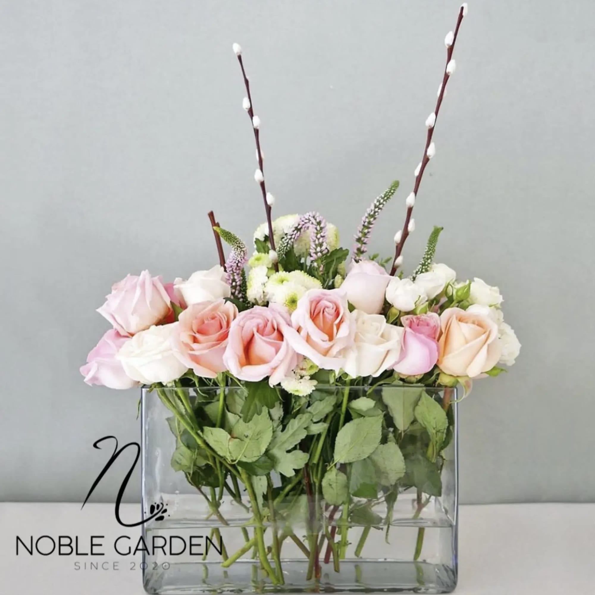 Pink and white roses in a clear rectangular vase with tall pussy willow stems