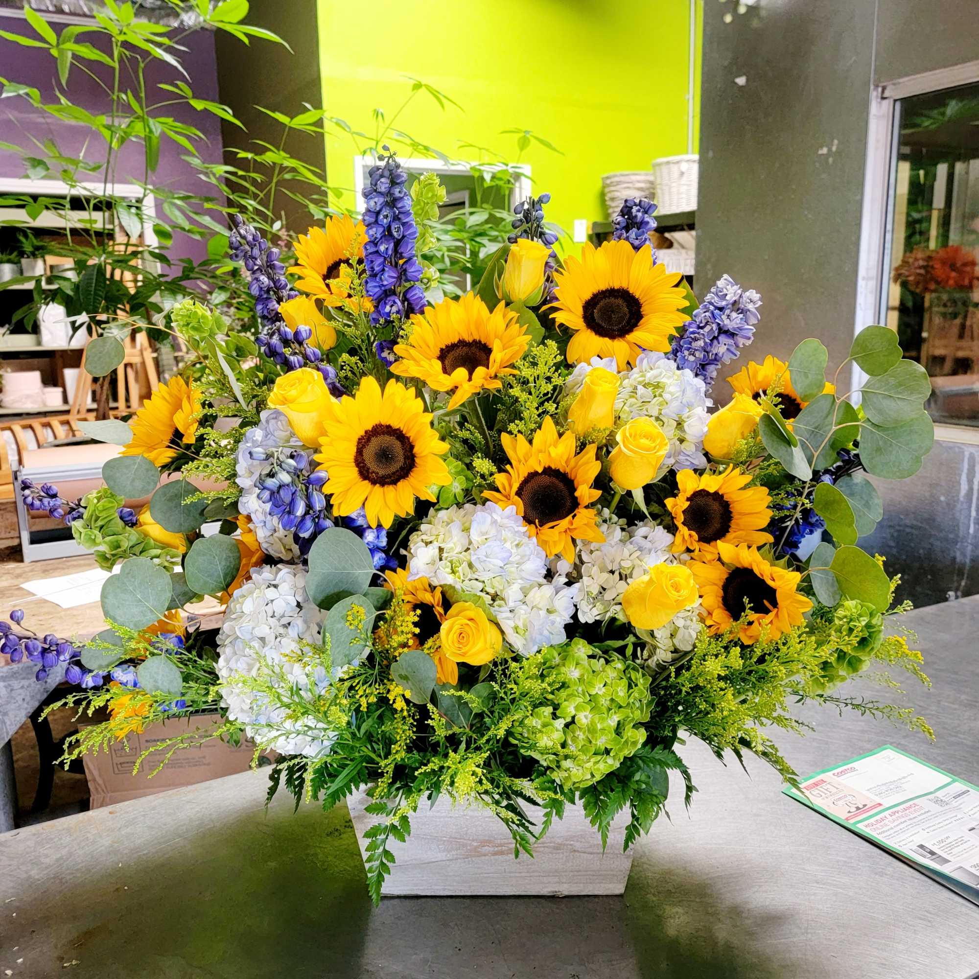 Sunflower arrangement with yellow roses, blue flowers, and white hydrangeas in a white container