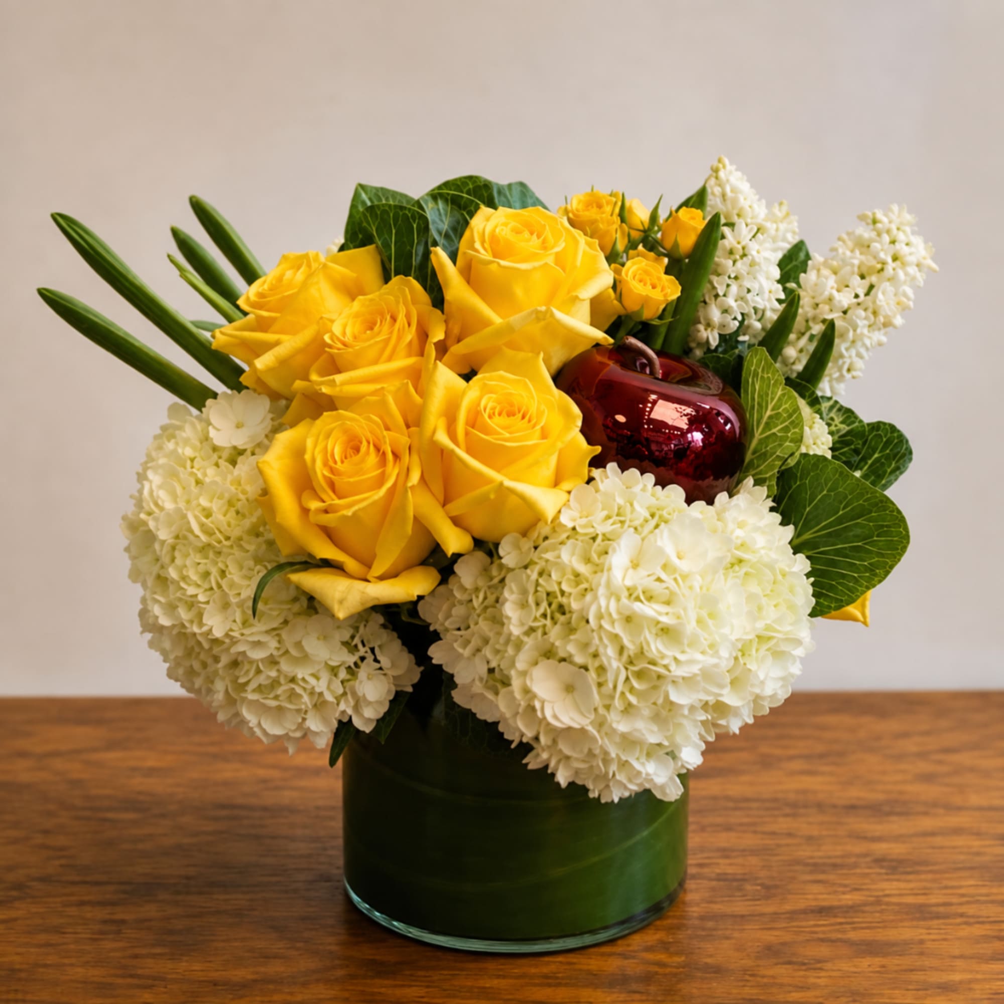 Yellow roses and white hydrangeas in a green glass vase with a red apple accent