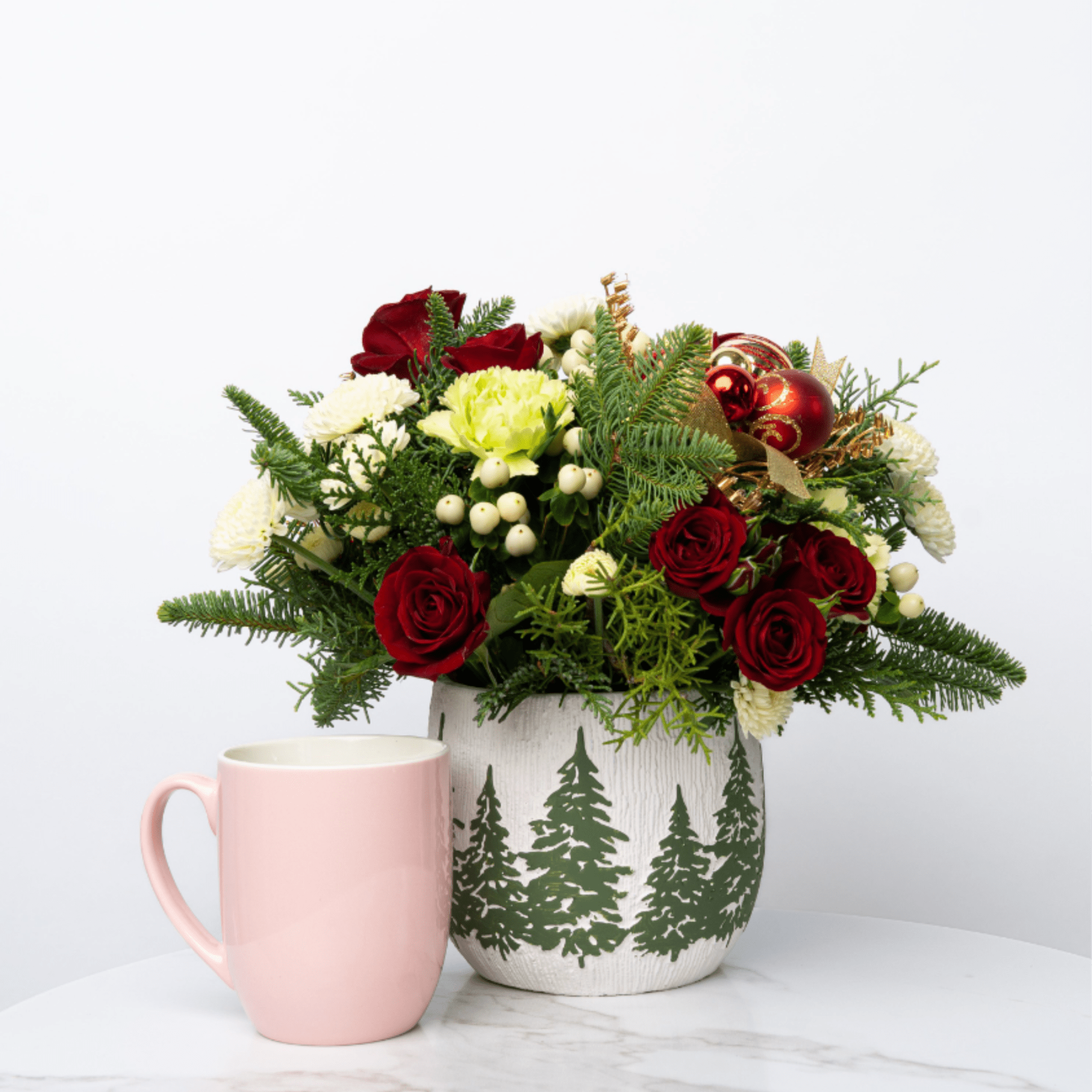 Red and white floral arrangement in a pine-decorated vase beside a pink mug