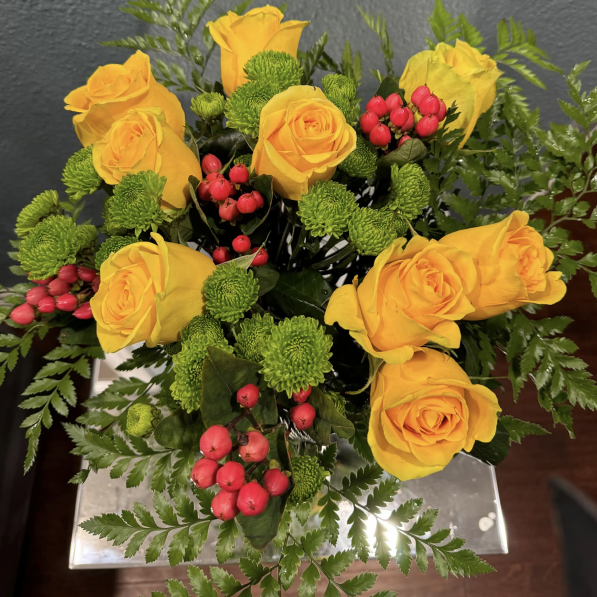 Yellow roses with green button mums and red berries in a clear vase