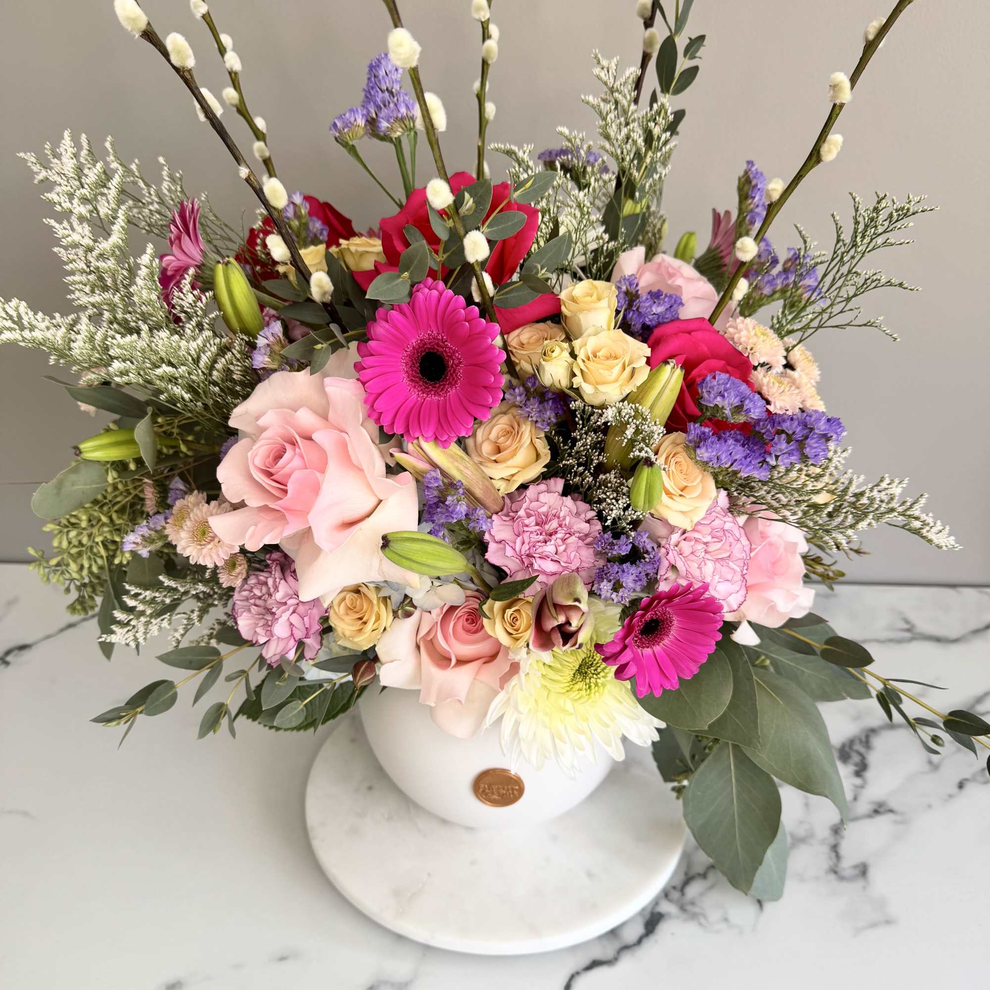 Colorful arrangement of pink gerberas, roses and mixed blooms in a white round vase with tall pussy willow branches