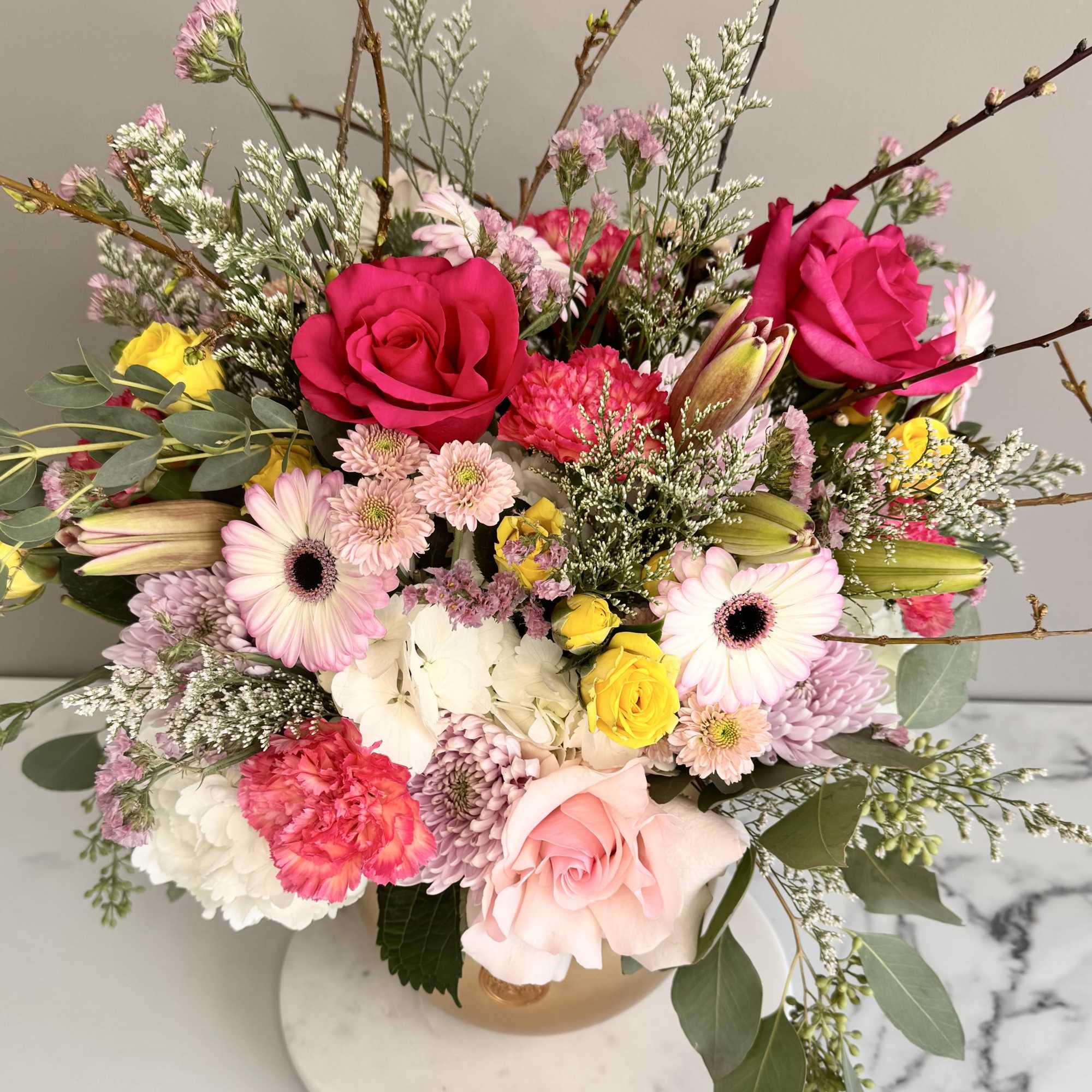 Lush pink, white, and yellow flower arrangement with roses, gerberas, and branches in a round vase
