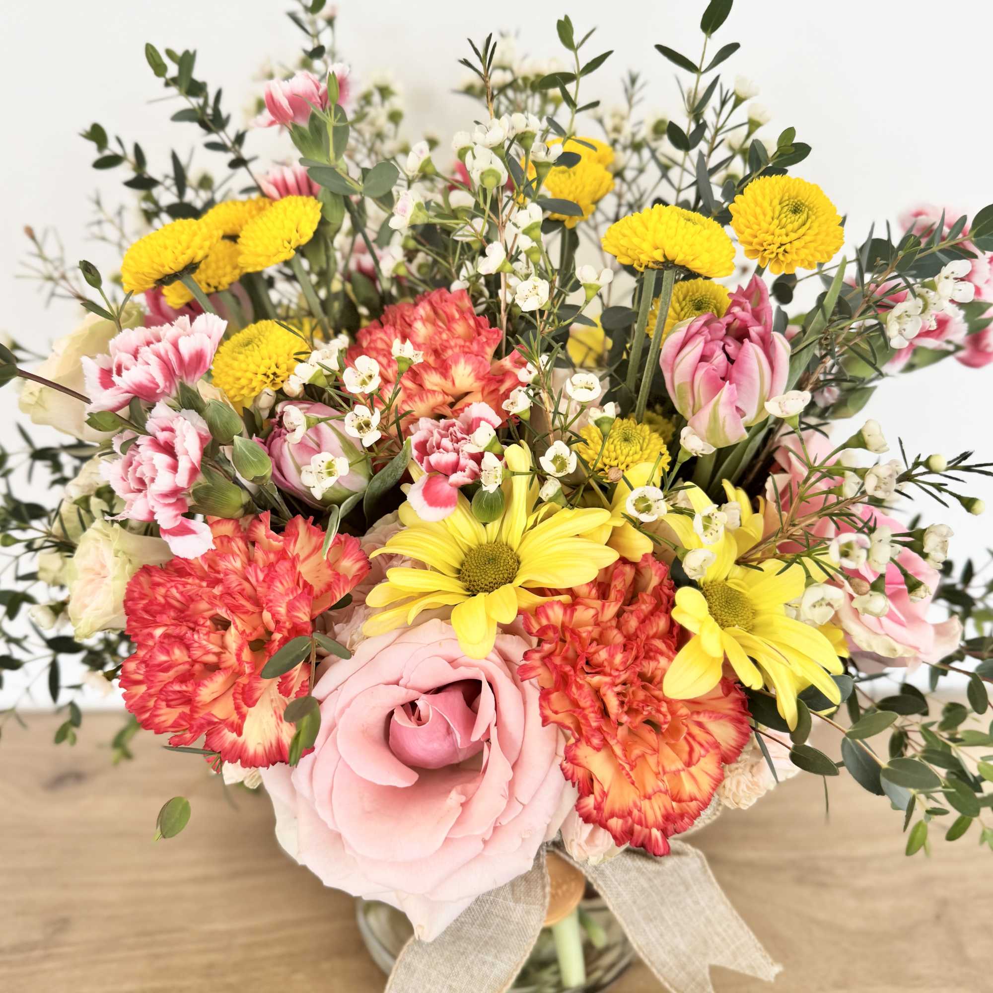 Cheerful bouquet of pink roses, orange carnations, and yellow daisies in a glass vase with a burlap bow.