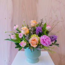 Pastel arrangement of peach roses, pink peony, and purple blooms in a light teal pedestal vase