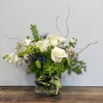 Low glass vase arrangement of white roses, white stock, green blooms, and blue thistle with a green ribbon.