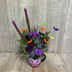 Purple floral arrangement in a smiling heart-shaped mug with butterfly picks