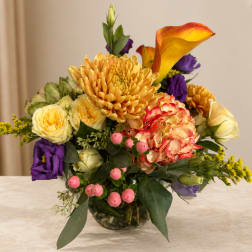 Mixed bouquet with yellow roses, orange calla lily, and purple blooms in a glass vase