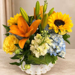Orange lilies, yellow sunflowers, roses, and blue hydrangeas in a white vase
