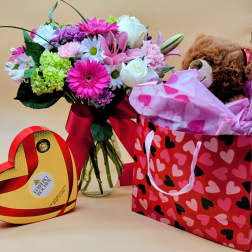 Mixed bouquet in a glass vase beside a teddy bear gift bag and heart-shaped chocolate box
