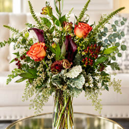 Tall bouquet of orange roses, dark calla lilies, and greenery in a glass vase