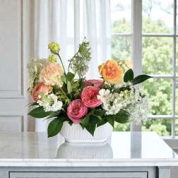 Low white bowl of pink, peach, and yellow flowers with white accents on a marble countertop.