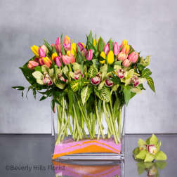 Rectangular vase of pink and yellow tulips with green orchids and variegated leaves over layered colored sand.