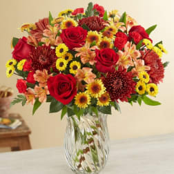 Mixed bouquet of red roses, yellow daisies, and orange flowers in a clear glass vase