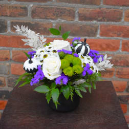 White roses and purple flowers arranged in a black vase with small decorative pumpkins