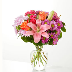 Pink lilies, orange roses, and purple daisies in a clear glass vase