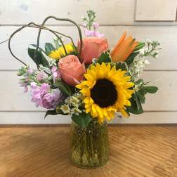 Mixed bouquet with sunflowers, peach roses, lilies, and lavender blooms in a green glass vase