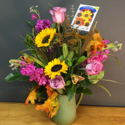 Mixed bouquet with sunflowers, pink roses, and purple blooms in a green pitcher vase