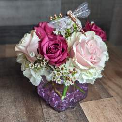 Short cube vase arrangement of pink and cream roses with white blooms and a decorative dragonfly pick.