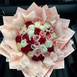 Round bouquet of red, pink, and white roses wrapped in layered peach paper with pearl accents.