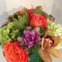 Compact arrangement of orange roses, green hydrangeas, purple flower, and tan orchid in a clear glass vase