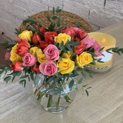 Round glass vase of red, pink, and yellow roses accented with eucalyptus on a light wood table.