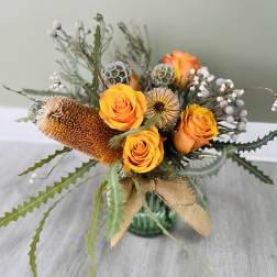 Orange roses in a glass vase with dried seed pods and a burlap ribbon