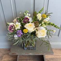 Mixed bouquet of cream roses, purple blooms, and yellow accents in a glass vase