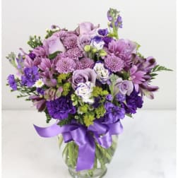Lavender and purple mixed flower arrangement with roses and mums in a clear vase with a satin ribbon bow