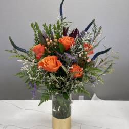 Orange roses in a clear glass vase with mixed greenery and purple accents