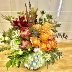 Mixed bouquet with orange roses, blue hydrangea, and burgundy protea in a low vase