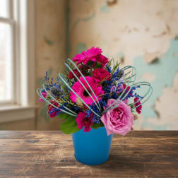 Bright pink gerbera daisies, rose, and carnations with blue accents in a blue pot.
